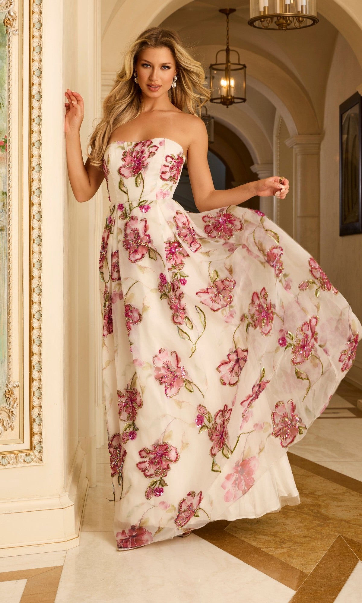 A woman wears the Long Prom Dress: Jovani 44645 in Off White/Multi, featuring a strapless floral ballgown design, as she stands in an ornate hallway holding her dress and smiling at the camera.